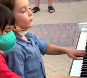 Two six-year-old boys are playing the piano right in the middle of the street. Passers-by gathered around them to listen to their performance.